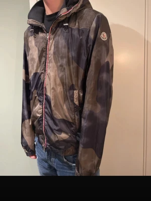 Moncler camo vindjacka med huva - Säljer en snygg vindjacka från Moncler med camouflagemönster i mörkblått och brunt. Jackan har huva, dragkedja med röd och blå detalj, samt Moncler-logga på ärmen. Perfekt för dig som gillar streetwear och vill ha något unikt. (5)