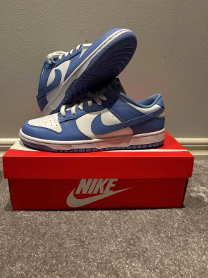 Nike Dunk Low blå och vita sneakers - Nike Dunk Low sneakers i en fräsch blå och vit färgkombination. Skorna har klassisk snörning, rund tå och platt sula. Ovandelen är tillverkad i skinn med perforerade detaljer på tån för extra ventilation. Snygg och stilren siluett som passar till streetwear.