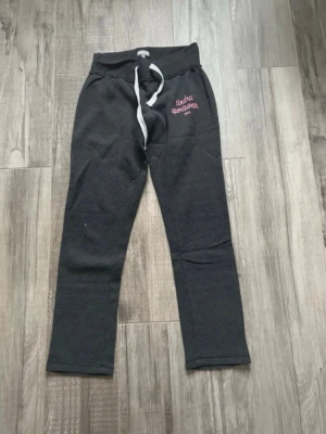 Mörkgrå mjukisbyxor från Tindra Sportswear - Grå mjukisbyxor från Tindra Sportswear med broderad rosa logga på vänster sida och vit snörning i midjan. Byxorna har normal passform och är tillverkade i mjukt bomullsmaterial. Perfekta för chill eller träning. Innerbenslängd 81 cm. Finns lite ytludd.