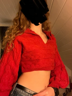 Röd croppad stickad tröja med krage - Croppad röd stickad tröja med kabelstickat mönster och bred krage. Tröjan har långa ärmar och en relaxed fit, perfekt för lager-på-lager. Snygg till både jeans och kjol för en trendig look.