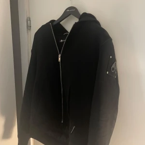 Svart hoodie/jacka från Moose Knuckles  - Svart hoodie från Moose Knuckles i barnstorlek xl ( jackan är väldigt liten i sin storlek så ja skulle säga att den passar någon som är runt 160-165). Snygg detalj med logotyp-patch på ärmen. Klassisk passform och mjukt material. Jackan är väldigt skön att ha på sig, passar perfekt när det är lite kyligt ute 