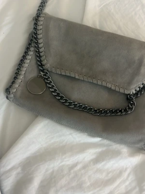 Grå axelväska med kedjedetaljer - Snygg grå axelväska i mockaimitation med grova kedjedetaljer i metall runt kanterna och som axelrem. I bra skick , kommer inte till användning. Pris kan diskuteras! 
