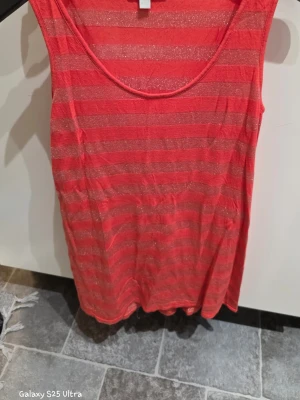 Röd glittrig randig linnetopp - Säljer en röd linnetopp från B.Young i storlek 40/42. Toppen har glittriga horisontella ränder och är ärmlös med rund halsringning. Materialet är mjukt och lätt, perfekt för varma dagar. Snygg att styla med jeans eller shorts för en färgstark look.