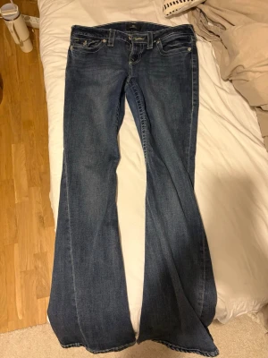 True Religion blå bootcut jeans - Säljer ett par klassiska blå jeans från True Religion med bootcut passform. Jeansen har kontrastsömmar, ikoniska fickdetaljer bak med knappar och en snygg tvätt. Tillverkade i slitstarkt denim med låg midja och femficksmodell.