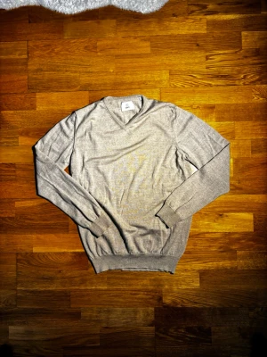 Beige v-ringad tröja från Bläck - Snygg beige v-ringad tröja från Bläck i storlek M. Tröjan har lång ärm, ribbade muddar och är gjord i ett mjukt stickat material som känns skönt mot huden. Perfekt för dig som gillar stilrena och enkla plagg med en klassisk look.