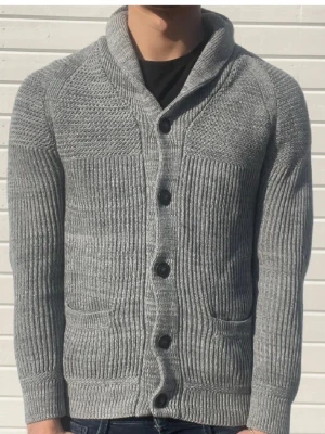 Grå stickad kofta från H&M - Säljer en grå stickad kofta från H&M med bred krage och fem knappar framtill. Koftan har två fickor och är ribbstickad med olika stickmönster på axlar och ärmar. Perfekt för kyliga dagar och enkel att matcha med jeans eller t-shirt.