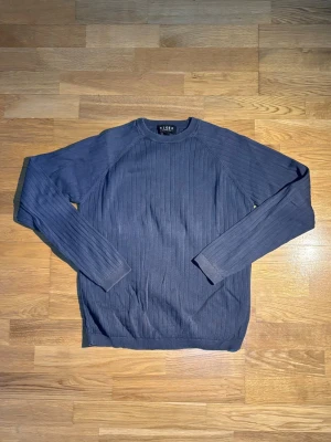 Mörkblå stickad tröja från Tiger - Säljer en mörkblå stickad tröja från Tiger med ribbad struktur och rund hals. Tröjan har långa ärmar och är perfekt för dig som gillar klassisk stil med en modern touch. Passar dig som vill ha en enkel men snygg look.