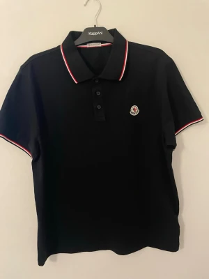 Svart Moncler piké med röd/vit kant  - Säljer en svart pikétröja från Moncler med klassisk krage och knäppning framtill. Pikén har röda och vita ränder på krage och ärmslut samt Moncler-logga broderad på bröstet. Perfekt för en clean och sportig look.