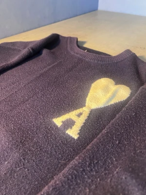 Mörklila stickad tröja från Ami - Snygg mörklila stickad tröja från Ami Alexandre Mattiussi med ett stort vitt hjärta och bokstaven A på bröstet. Tröjan har rund hals, ribbade muddar och långa ärmar. Perfekt för dig som gillar stilrena detaljer och vill ha något extra i garderoben.
