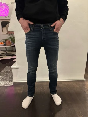 Mörkblå skinny jeans från Jack & Jones - Säljer ett par mörkblå skinny jeans från Jack & Jones med snygga slitningar och klassisk femficksdesign. Jeansen har låg midja och smal passform som sitter tajt längs benen. Materialet är stretchigt jeans och de har en knapp och dragkedja framtill. Modellen är 175 cm, vikt 68 kg