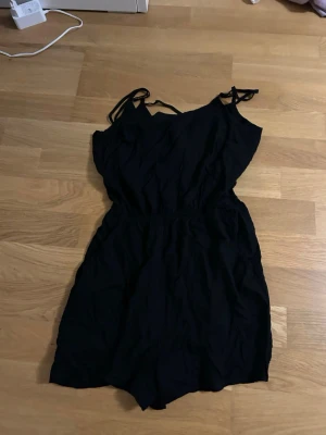 Svart byxdress från Divided strl 32 - Svart byxdress från Divided i storlek 32. Modellen har smala axelband med knyt och resår i midjan för en skön passform. Materialet är mjukt och lätt bomull, perfekt för varma dagar. Enkel och stilren design med korta ben och omlott-detalj upptill.