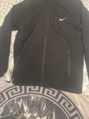 Svart vindjacka från Nike - Säljer en svart vindjacka från Nike med klassisk logga på bröstet. Jackan har hel dragkedja framtill och två sidofickor med dragkedja. Perfekt för dig som gillar sportig stil och vill ha något lätt och snyggt att slänga på sig.