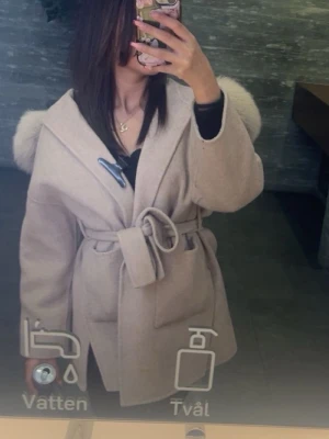 Beige kappa med pälsdetaljer och bälte - Säljer en beige kappa med stora fluffiga pälsdetaljer på axlarna och breda slag. Jackan har ett matchande bälte i midjan och en snygg omlottstil. Perfekt för dig som gillar en chill och lyxig vibe. Långärmad och med fickor framtill.