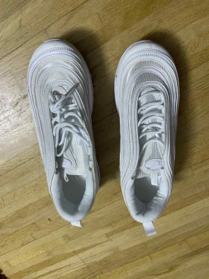 Nike Air Max 97 helvita sneakers - Nike Air Max 97 sneakers i helvit färg med ikoniska vågiga linjer och synlig Air-sula. Skorna har snörning, rund tå och platt sula. Ovandelen är i syntet och mesh med broderad Nike-logga på sidan. Perfekt för dig som gillar clean och sportig stil.