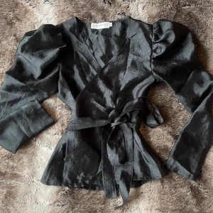 Svart omlottblus med puffärm - Säljer en svart omlottblus från IDA SJÖSTEDT med puffiga axlar och knytband i midjan. Blusen har lång ärm och glansigt material som ger en lyxig känsla. Perfekt för dig som vill ha en statement-top med snygga detaljer.❤️