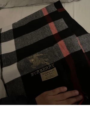Rutig Burberry halsduk 100% Kashmir  - Säljer en riktigt snygg rutig halsduk  från Burberry i 100% kashmir. Västen har klassiskt Burberry-mönster i svart, grått, vitt och röda detaljer samt broderad logga och etikett framtill. Perfekt för dig som vill ha något stilrent och lyxigt.