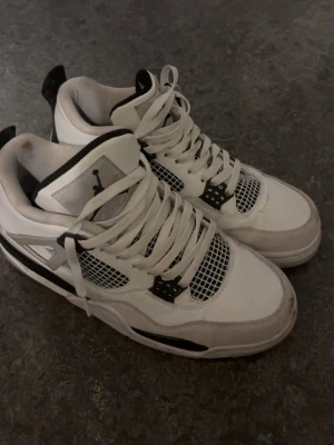 Nike Air Jordan 4 vit/svart - Säljer ett par Nike Air Jordan 4 i vit och svart med klassisk meshpanel, snörning och ikoniska Jumpman-loggan på hälen. Skorna har en chunky sula och detaljer i mocka och skinn för en riktigt clean och sportig look.