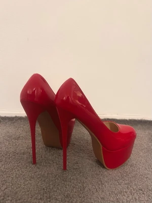 Röda pumps med hög klack och plattform - Säljer ett par snygga röda pumps med riktigt hög klack och plattform. Skorna har rund tå och är tillverkade i glansigt syntetmaterial som ger en cool och trendig look. Perfekta för dig som vill sticka ut och älskar färg!