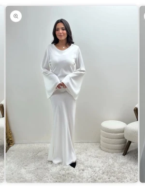 Vit långklänning med vida ärmar - Elegant vit långklänning med vida ärmar och snörning i ryggen. Klänningen har en mjuk, flytande passform och är tillverkad i ett glansigt material som ger en lyxig känsla. Perfekt för dig som vill sticka ut med en stilren och modern look. Beställde klänningen från Paris och aldrig använd för ville returnera då jag inte behövde den  längre men hemsidan står ej för frakten så gjorde ej det