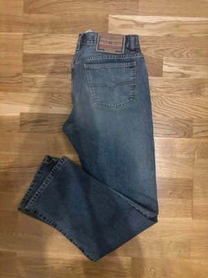 Diesel jeans  - Snygga blå jeans från Diesel i klassisk straight fit. Modellen har fem fickor, knappgylf och coola detaljer som bruna sömmar och läderpatch bak. Tillverkade i Italien av slitstarkt jeanstyg som ger en autentisk känsla. Perfekt för dig som gillar tidlös denimstil.