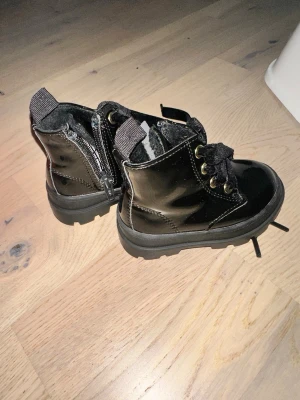 Svarta boots med snörning från H&M H&M Svarta Boots Stl 18 - Säljer ett par svarta boots från H&M med grov platt sula och snörning i sammet. Skorna har dragkedja på sidan för enkel på- och avtagning samt detaljer i guld vid snörningen. Yttermaterialet är blankt syntet och insidan är fodrad med mjukt tyg.Ett par svarta boots från H&M. Skorna har snörning framtill och dragkedja på sidan.