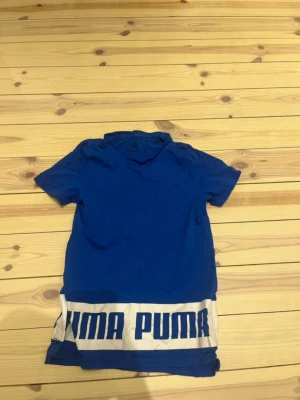 Blå Puma t-shirt med tryck - Blå t-shirt från Puma med korta ärmar och stort vitt tryck med blå text 'PUMA' längs nederkanten. Materialet är bomull och polyester, perfekt för träning. Diskret Puma-logga i vitt på ryggen och insidan har texten 'Keeps You Dry'. Hör av er vid frågor 