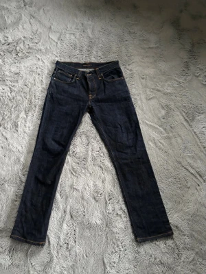 Grim Tim från  Nudie 32x32 - Snygga mörkblå jeans från Nudie med klassisk femficksdesign och kontrastsömmar i orange. Modellen har rak passform och normal midja, tillverkade i slitstarkt bomullsmaterial. Perfekta för dig som gillar stilrena och tidlösa jeans.