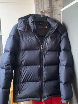 Svart pufferjacka från Polo Ralph Lauren - Svart pufferjacka från Polo Ralph Lauren med huva och två sidofickor med dragkedja. Jackan har en röd broderad logga på bröstet och RL-detalj på dragkedjan. Perfekt för kalla dagar och har en snygg, sportig look. ”Möts upp i Stockholm mvh , noel😁