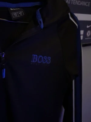 Svart hoodie från BOSS med blå/vita detaljer - Säljer en svart hoodie från BOSS i storlek 176, med blå och vita ränder längs ärmarna och BOSS-logga på bröstet. Tröjan har huva, dragkedja framtill och fickor med dragkedja. Perfekt för dig som vill ha en clean och sportig look.