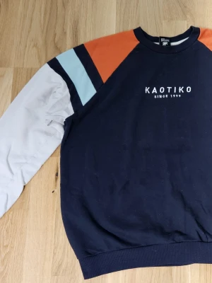 Mörkblå sweatshirt från Kaotiko - Sweatshirt från Kaotiko i mörkblått med vita, ljusblå och röd/orange detaljer på axlar och ärmar. Tröjan har långärmad design och ribbade muddar vid hals och ärmslut. Tryck med 'KAOTIKO SINCE 1999' på bröstet. Perfekt för chill och streetwear. Riktigt skön att ha på sig.