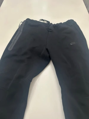 Svarta mjukisbyxor från Nike XXL - Svarta mjukisbyxor från Nike i storlek XXL. Byxorna har dragsko i midjan, ribbade muddar vid bensluten och en diskret Nike-logga på benet. Materialet är mjukt och bekvämt, perfekt för chill eller träning. Snygga detaljer med dragkedja på fickan.