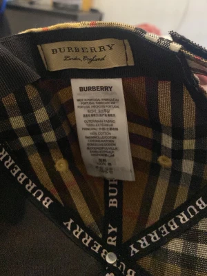 Rutig Burberry keps i bomull - Säljer en klassisk Burberry keps i 100% bomull med det ikoniska rutiga mönstret i beige, svart, rött och vitt. Kepsen har böjd skärm och justerbar passform. Perfekt accessoar för att lyfta din outfit med en lyxig touch.