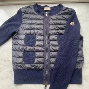Moncler cardigan  - Snygg mörkblå cardigan från Moncler med quiltad front och stickade ärmar. Jackan har två stickade fickor fram, ribbade muddar och dragkedja. Perfekt för dig som vill ha en lätt och stilren jacka med exklusiv känsla.