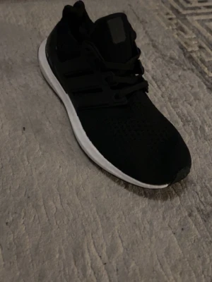 Adidas Ultraboost svarta sneakers - Svarta Adidas Ultraboost sneakers med vit sula och klassiska tre ränder på sidan. Ovandelen är i ventilerande syntetmaterial och skon har snörning. Perfekt för dig som gillar stilrena och sportiga skor med skön komfort.