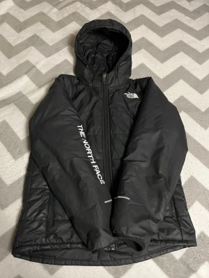 Svart pufferjacka från The North Face pris kan diskuteras  - Svart pufferjacka med huva från The North Face. Jackan har dragkedja framtill, vita logotryck på bröstet och ärmen, samt två sidofickor med dragkedja. Materialet är syntet och jackan har en quiltad design på ryggen. Perfekt för kyliga dagar.
