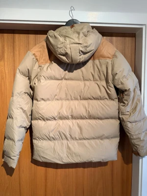 Peak performance Beige Dun jacka  M - Säljer en beige pufferjacka från Peak Performance i storlek M. Jackan har bruna detaljer på axlarna, huva med snörning och flera fickor framtill. Materialet är polyester och jackan är perfekt för kalla dagar. Snygg och funktionell design med dragkedja och knappar.
