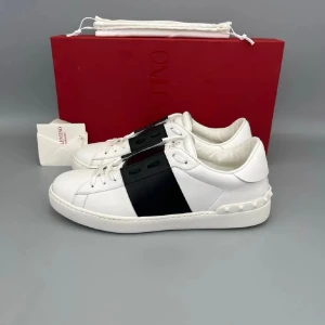 Valentino Garavani Open sneakers vit/blå - Snygga Valentino Garavani Open sneakers i vitt skinn med bred svart rem över mitten. Klassisk rund tå, platt sula och vita nitar längs hälen ger en lyxig touch. Skorna har snörning och logga på plösen. Perfekt för dig som gillar stilrena designer.