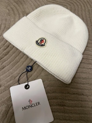 Vit Moncler mössa i ull - Säljer en stilren vit Moncler mössa i ribbstickad ull. Klassisk uppvikt kant och ikoniska Moncler-loggan framtill. Perfekt accessoar för kalla dagar och snygg till alla outfits.