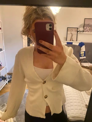 Vit kofta från Gina Tricot med knappar - Säljer en stilren vit kofta från Gina Tricot i storlek M. Koftan har v-ringad hals, stora dekorativa knappar framtill och på ärmsluten. Modellen är figurnära med lång ärm och mjukt stickat material som känns skönt mot huden.