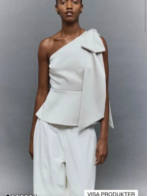 Vit oneshoulder topp med stor rosett - Supersnygg vit topp i satin med oneshoulder-design och en stor rosett på axeln. Toppen har figurnära passform och är croppad med lätt utsvängd nederkant. Perfekt för dig som vill sticka ut med en elegant och trendig look.