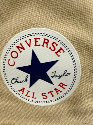 Beige Converse Chuck Taylor All Star - Säljer ett par klassiska Converse Chuck Taylor All Star high tops i beige canvas med vita gummisulor och tå. Skorna har vit snörning, rund tå och den ikoniska loggan på sidan. Perfekt för dig som gillar retrostil och vill ha ett par tidlösa sneakers.