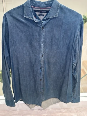 Blå jeansskjorta Tommy Hilfiger - Snygg blå jeansskjorta från Tommy Hilfiger i storlek L. Skjortan har klassisk krage, långa ärmar och knappar framtill. Tillverkad i mjuk bomull som ger en avslappnad känsla. Perfekt för dig som gillar stilren denim och vill ha en enkel look.