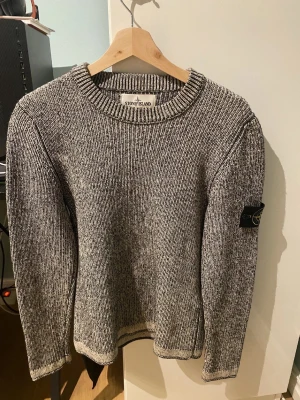 Stickad Stone Island tröja (äkta) - Säljer en snygg gråmelerad stickad tröja från Stone Island med rund hals och lång ärm. Tröjan har en svart logotyp-patch på vänster ärm och ribbade muddar vid hals, ärmslut och nederkant. Perfekt för kyliga dagar och enkel att matcha med jeans. Priset är inte helt satt så passa på och skicka pris förslag. Den är M men har krympt till S