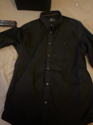 Svart skjorta från Madalyon slim fit - Svart skjorta från Madalyon i slim fit-modell, med klassisk krage och knappar hela vägen framtill. Skjortan har lång ärm och är tillverkad i ett mjukt bomullstyg. Perfekt för dig som gillar stilrena och enkla plagg.