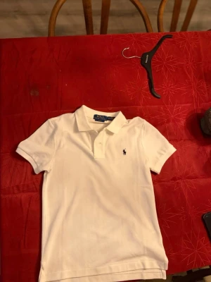 Vit piké från Polo Ralph Lauren - Säljer en klassisk vit pikétröja från Polo Ralph Lauren i slim fit-modell. Tröjan har korta ärmar, krage och två knappar framtill. Ikoniska loggan broderad på bröstet. Perfekt för en clean och stilren look.