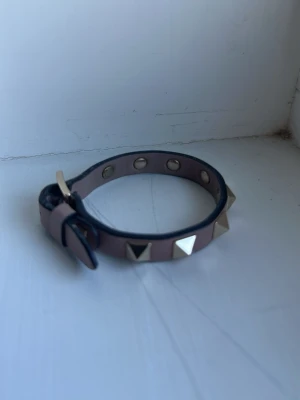 Rosa skinnarmband med nitar - Säljer ett snyggt armband i rosa skinn med silverfärgade pyramidnitar och spänne. Perfekt accessoar för att ge din outfit lite edge. Justerbar storlek med flera hål för passform. Har liten defekt men syns inte när man sätter fast armbandet.