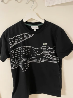 Svart t-shirt med krokodil  Lacoste - Cool svart t-shirt från Lacoste med ett stort grafiskt krokodilmotiv i vitt och grått över framsidan. Klassisk rund hals och korta ärmar. Materialet är mjuk bomull och den har den ikoniska gröna Lacoste-loggan på bröstet.Liknande modell som originala.