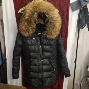 Svart pufferjacka Rockandblue med päls - Svart pufferjacka från Rockandblue med stor fluffig fuskpäls runt huvan. Jackan har quiltad design, dragkedja och knappar framtill samt flera fickor med dragkedja. Perfekt för kalla dagar och har en riktigt snygg siluett. Den har även 2 st innerfickor och en extra knapp på insidan. Mått : Axel till Axel : ca 43 cm. Arm håla till Armhåla :ca 48 cm. Armlängd: ca 60 cm. Längd ( uppifrån ,,ner): ca 73 cm . 