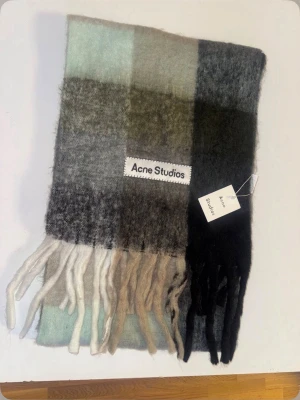 Rutig Acne Studios ullhalsduk - Säljer en stor och fluffig halsduk från Acne Studios i rutigt mönster. Färgerna går i svart, grått, beige och ljusblått. Halsduken har långa fransar i ändarna och en stor Acne Studios-logga. Materialet är mjuk ull som känns lyxigt och värmande.