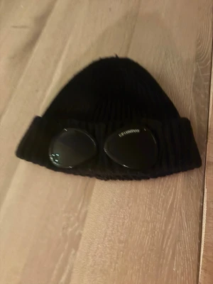 Svart mössa med glasögon från C.P. Company - Säljer en svart ribbstickad mössa från C.P. Company med inbyggda glasögon på uppviket. Mössan har en cool och unik design som sticker ut och är perfekt för dig som vill ha något annorlunda. Materialet är mjukt och bekvämt. Ny pris 1500